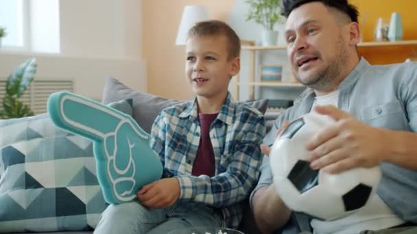 Homme joyeux et les petits garçons fans de sport profiter d'un match à la télévision tenant ballon sur le canapé à la maison — Video