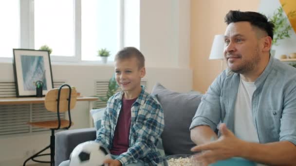 Papa en zoon kijken naar voetbal op tv vieren overwinning gooien bal hebben plezier thuis — Stockvideo