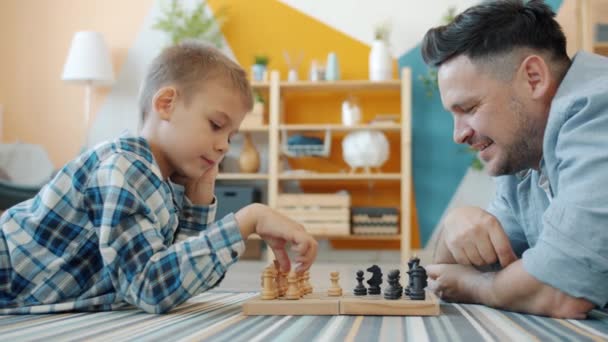 Niño inteligente jugando ajedrez con papá en casa tumbados en el suelo juntos divirtiéndose — Vídeo de stock