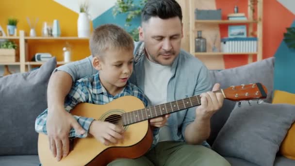 Liebenswerter Junge spielt Gitarre unter Anleitung des fürsorglichen Vaters, der zu Hause lernt — Stockvideo