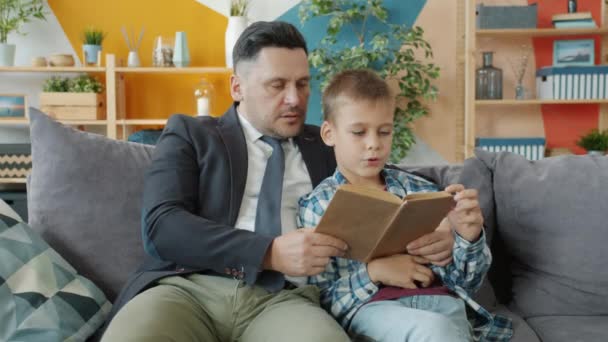 Vader in formele kleding lezen boek aan vrolijke kleine jongen in appartement — Stockvideo