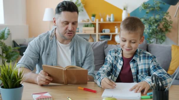 Bambino creativo che disegna immagini mentre il padre legge il libro seduto alla scrivania insieme — Video Stock