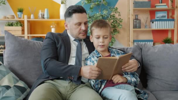 Tata biznesmen uczy małego syna czytać książkę trzymając siedzi na kanapie w domu — Wideo stockowe