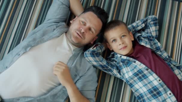 Retrato de hombre adulto y chico lindo tumbado en el piso de la casa hablando relajándose juntos — Vídeos de Stock