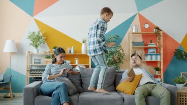 Hijo saltando en el sofá, mientras que la madre usando el teléfono inteligente padre libro de lectura haciendo high-five — Vídeo de stock