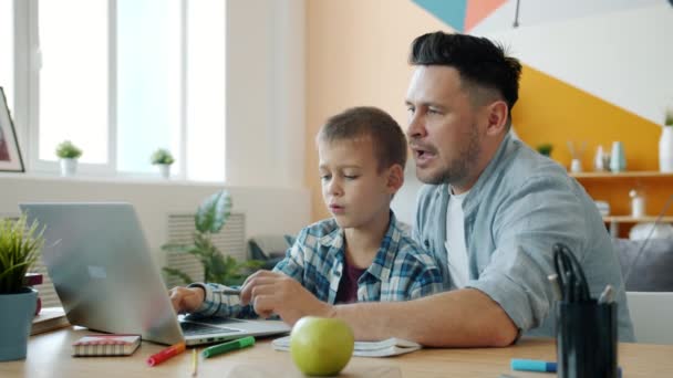 Kleine jongen leren online met behulp van laptop, terwijl vader helpt zitten aan tafel in huis — Stockvideo