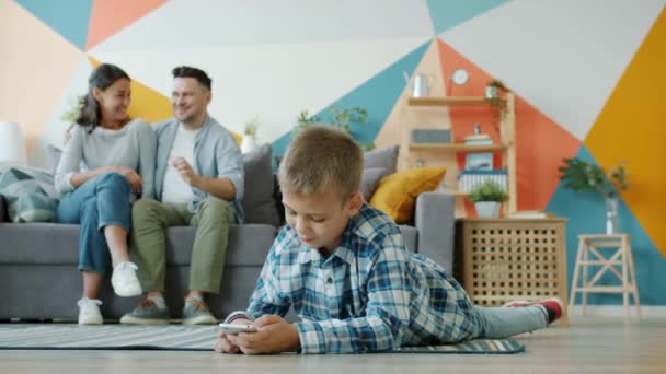 Повільний рух щасливого хлопчика за допомогою смартфона, коли батьки розмовляють на задньому плані — стокове відео