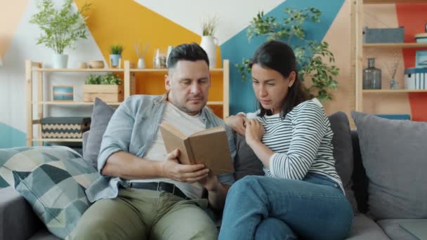 Молода пара чоловік і жінка читають книгу, розмовляючи, насолоджуючись літературою в квартирі — стокове відео