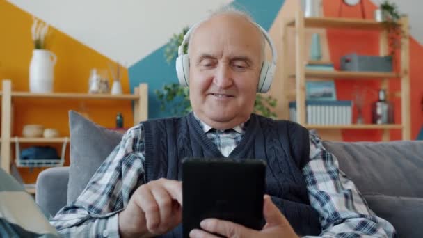 Hombre mayor sonriente en auriculares disfrutando de la música y utilizando la pantalla táctil de la tableta — Vídeos de Stock