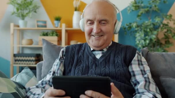 Gelukkige oudere man in hoofdtelefoon lachen tablet scherm met plezier thuis — Stockvideo