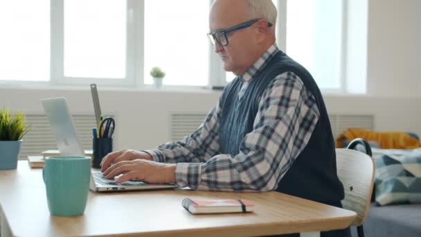 Vážné starší muž pomocí počítače doma psaní sedí za stolem uvnitř — Stock video