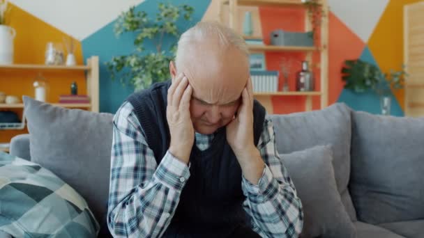 Starý muž trpí bolestí hlavy sedí na gauči sám dotýká nemocné hlavy — Stock video