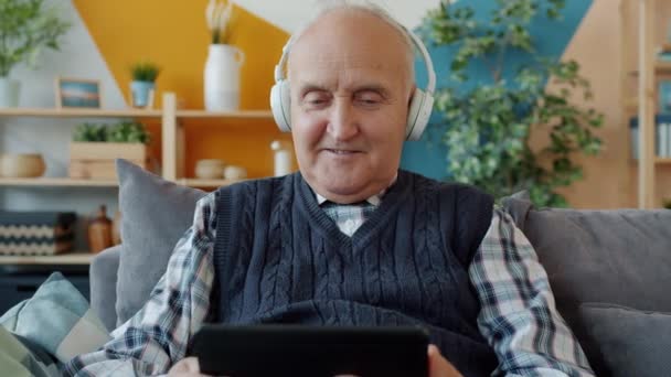 Симпатичный старик смотрит на экран планшета в наушниках в доме — стоковое видео