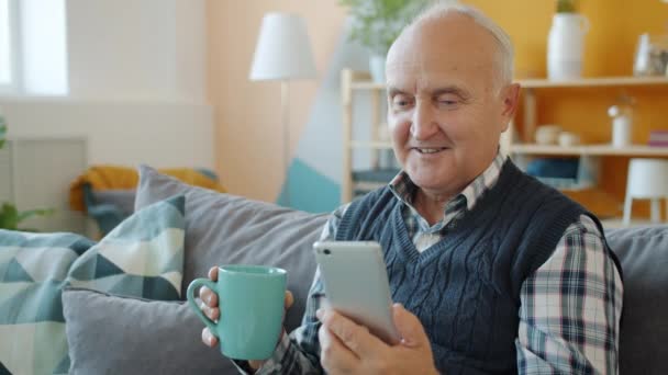 Älterer Mann schaut sich Inhalte auf Smartphone-Bildschirm an und trinkt zu Hause entspannt Tee — Stockvideo