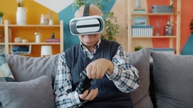 Üst düzey bir adam sanal gerçeklik gözlüğü ve joystick kullanıyor. Evdeki koltukta oturuyor.