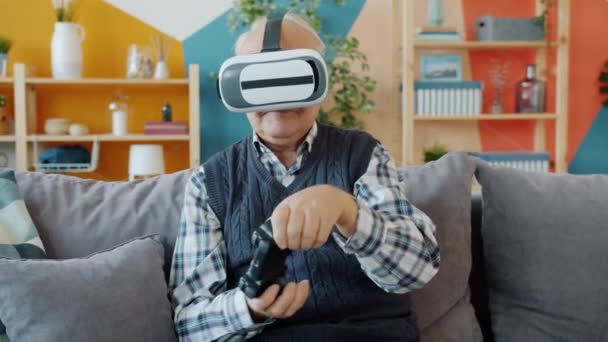 Hombre mayor usando gafas de realidad virtual y joystick sentado en el sofá en el apartamento — Vídeo de stock