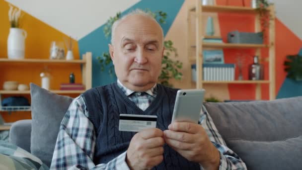 Happy old man making online payment with smartphone and credit card at home — Stock Video