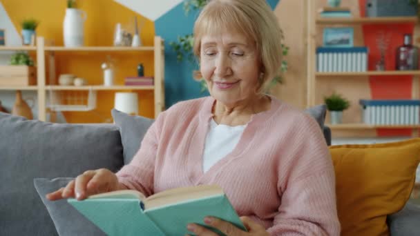 Mujer mayor sonriente leyendo libro sentado en el sofá en casa disfrutando de la novela — Vídeo de stock