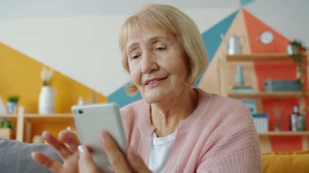 Медленное движение веселой женщины на пенсии с помощью смартфона трогательный экран улыбаясь дома — стоковое видео