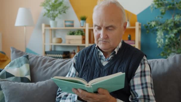 Homme âgé lisant le livre avec le visage sérieux tournant les pages se détendre sur le canapé à l'intérieur — Video