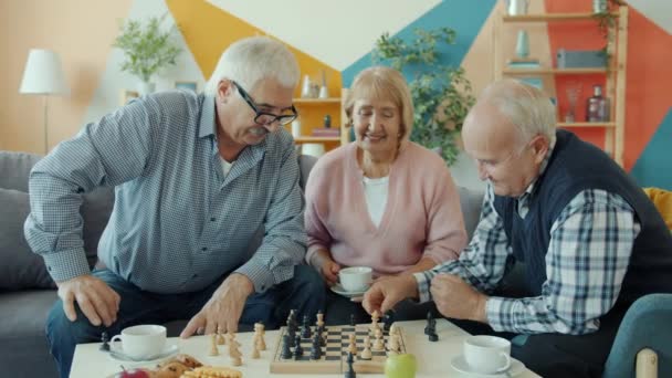 Les hommes âgés s'amusent avec le jeu d'échecs tandis que la vieille dame regarde et boit du thé à la maison — Video