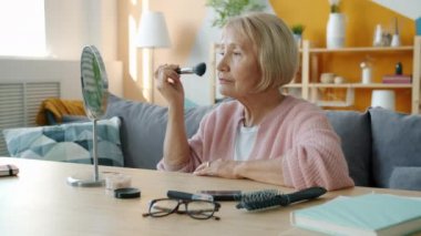 Çekici emekli kadın yüzünü güzelleştiriyor ve saçlarını tarıyor. Sonra da evde aynada gülümsüyor.