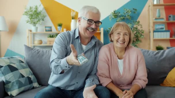 Πορτρέτο του ευτυχισμένου ηλικιωμένου ζευγαριού βλέποντας τηλεόραση στο σπίτι κρατώντας τηλεχειριστήριο απολαμβάνοντας αστεία ταινία — Αρχείο Βίντεο