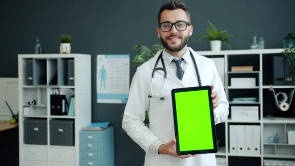 Wesoły młody człowiek lekarz w białej sukni trzyma tabletkę z zielonym ekranem chroma key uśmiechnięty — Wideo stockowe