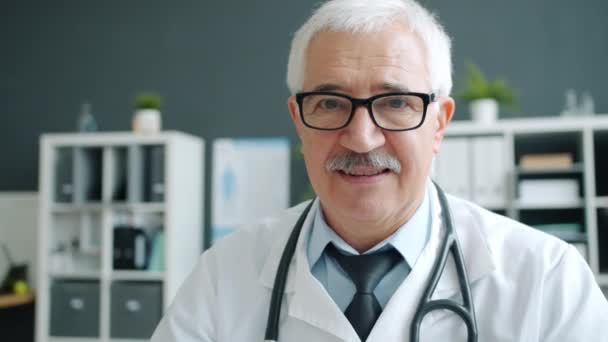 Medico anziano in uniforme e occhiali sorridenti in ufficio in attesa di pazienti in clinica — Video Stock