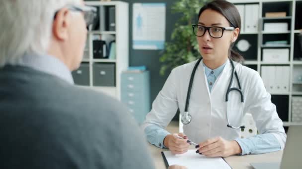 Mujer joven médico estrechando la mano con anciano paciente hombre hablando en el consultorio — Vídeos de Stock
