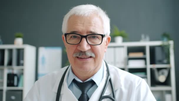 Portret siwowłosego lekarza w białym mundurze uśmiechniętego samotnie w biurze — Wideo stockowe