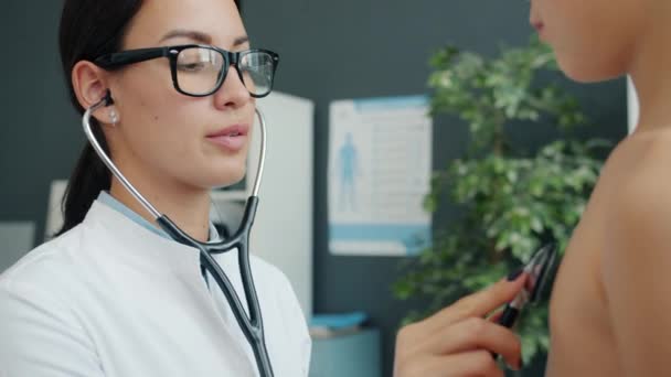 Young woman doctor examining cute little boy with stethoscope in office checking heartbeat — Stock Video