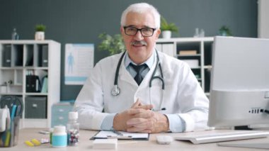 Beyaz önlüklü, gözlüklü gülümseyen doktor masada oturan kameraya bakıyor.