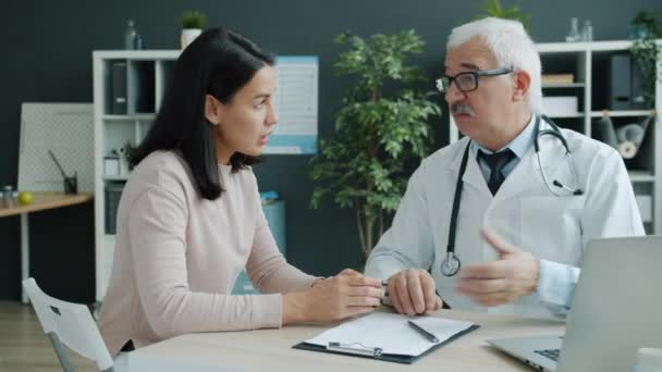 Médico cuidador hablando con una joven que toma de la mano expresando apoyo y cuidado en el consultorio — Vídeo de stock