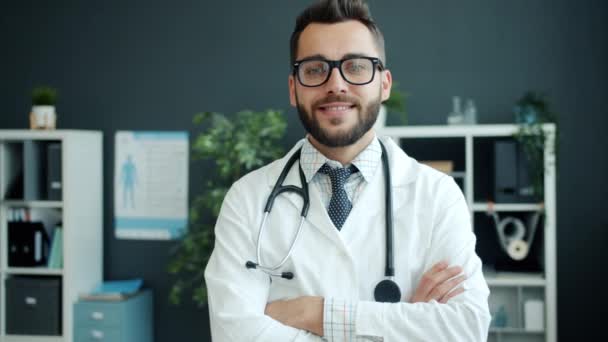 친절 한 젊은 의사가 두 팔을 꼬고 사무실에 서서 웃고 있는 모습 — 비디오