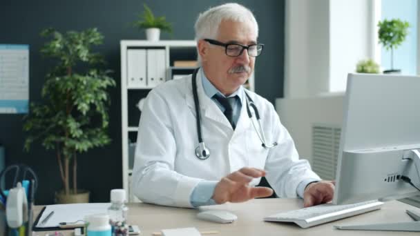 Vážný serior muž lékař pomocí počítače psaní sedí na stole v nemocniční kanceláři — Stock video