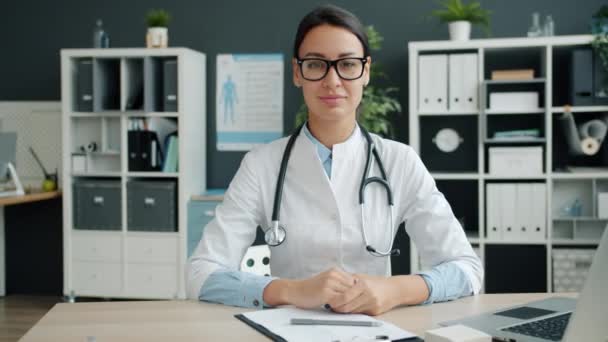 Portret van vrouwelijke arts kijkend naar camera met vriendelijke glimlach zittend aan bureau in ziekenhuiskantoor — Stockvideo