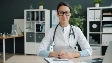 Beyaz önlüklü çekici genç kadın doktor portresi ofiste masada oturuyor.