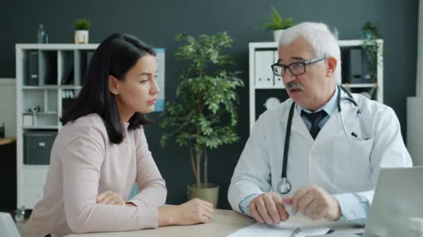Добрый старший врач разговаривает с пациентом, держащимся за руку, глядя на экран компьютера в офисе — стоковое видео