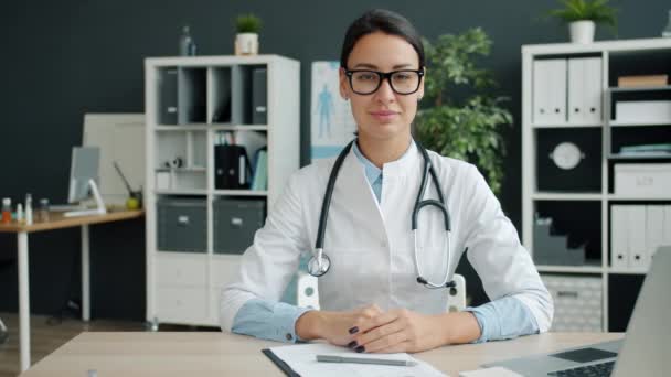 Retrato de atraente jovem médico em vestido branco sentado na mesa no escritório — Vídeo de Stock