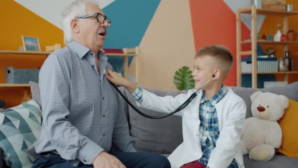 Oude man speelt met kind die hem onderzoekt met stethoscoop in huis — Stockvideo