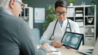 Genç bayan doktor, hastane ofisindeki yaşlı hastaya MRI sonuçlarıyla tablet ekranı gösteriyor.