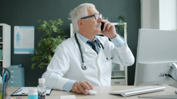 携帯電話で話すシニア医師と病院のオフィスルームでコンピュータを使用しています — ストック動画