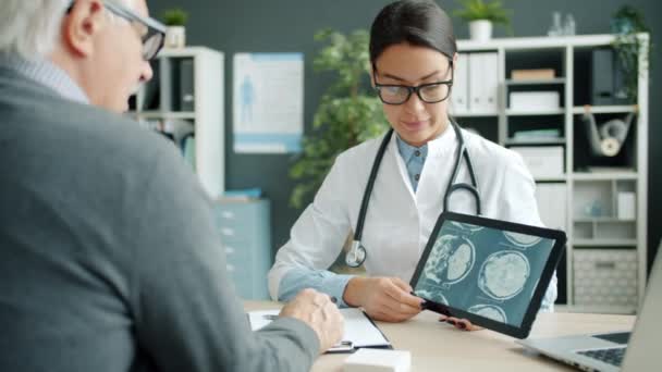 Genç bayan doktor, hastane ofisindeki yaşlı hastaya MRI sonuçlarıyla tablet ekranı gösteriyor. — Stok video