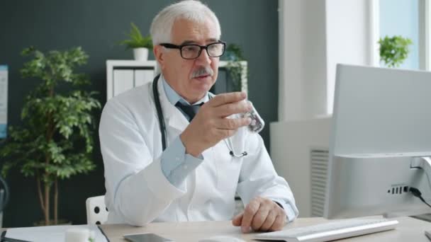 Médico en uniforme que tiene consulta en línea que habla mirando la pantalla del ordenador que muestra píldoras — Vídeo de stock