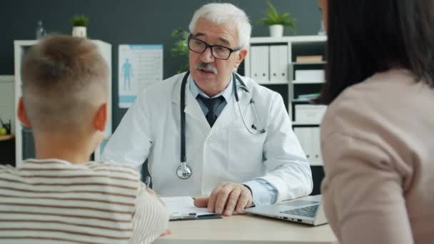 Pediatra con experiencia hablando con la madre y el niño escribiendo en el historial médico en la clínica — Vídeos de Stock