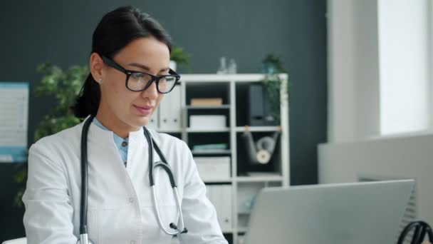 Χαμογελώντας νεαρή γυναίκα γιατρός χρησιμοποιώντας φορητό υπολογιστή στο χώρο εργασίας, στη συνέχεια, αγγίζοντας γυαλιά — Αρχείο Βίντεο