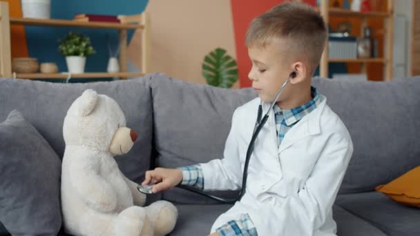 Enfant heureux en robe médicale blanche examinant ours en peluche avec stéthoscope à la maison — Video