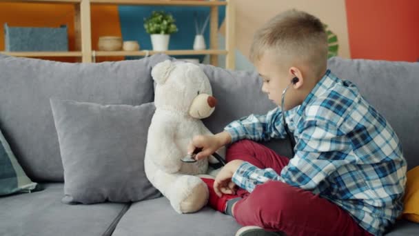 Милый ребенок играет в доктора с плюшевым мишкой, используя стетоскоп, улыбаясь дома — стоковое видео