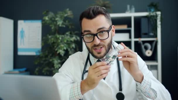 Портрет доброзичливого молодого лікаря, який показує таблетки і говорить, дивлячись на камеру в клініці — стокове відео